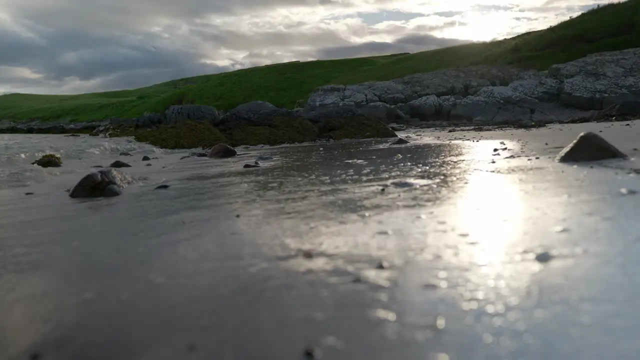 A low angle shot of waves gently lapping up against a sandy beach very close to the camera as the sun sets behind grassy hills in the background