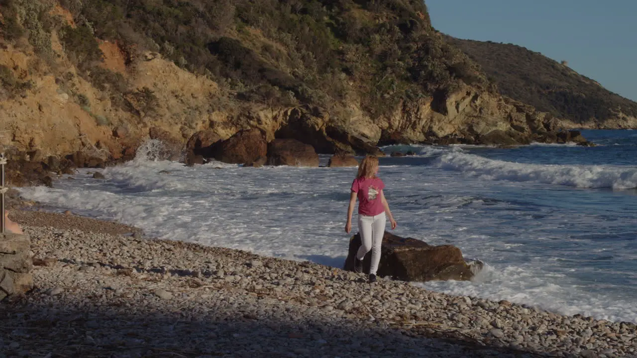 Girl walking on beach picks up a stone from ground strong waves crash sunny day