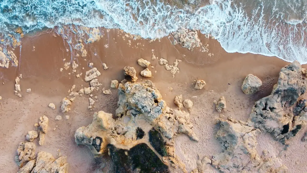 Top-down drone movement shot of rocky beach with waves gentle breaking into the sand