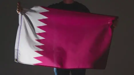 Studio Shot Of Anonymous Person Or Sports Fan Waving Flag Of Qatar Against Black Background