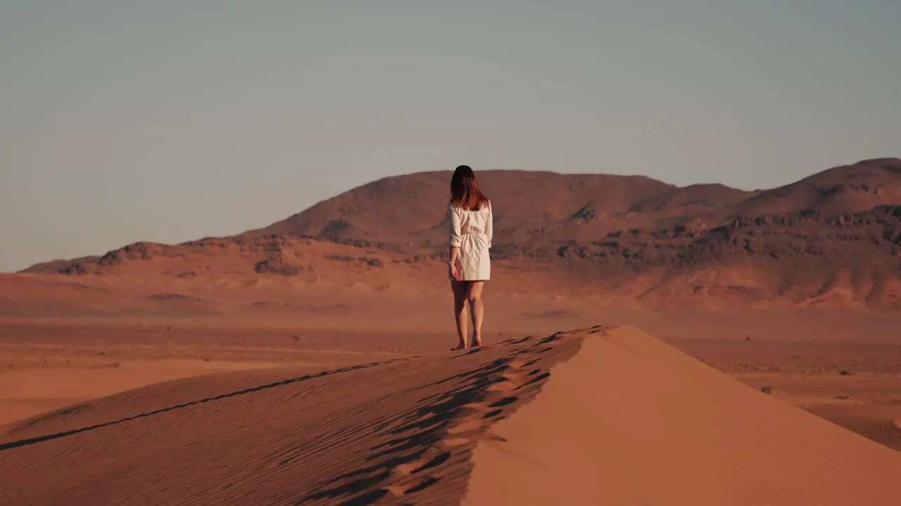 Young caucasian woman walking on top of a desert dune during sunset with mountains in the background in Zagora Morocco