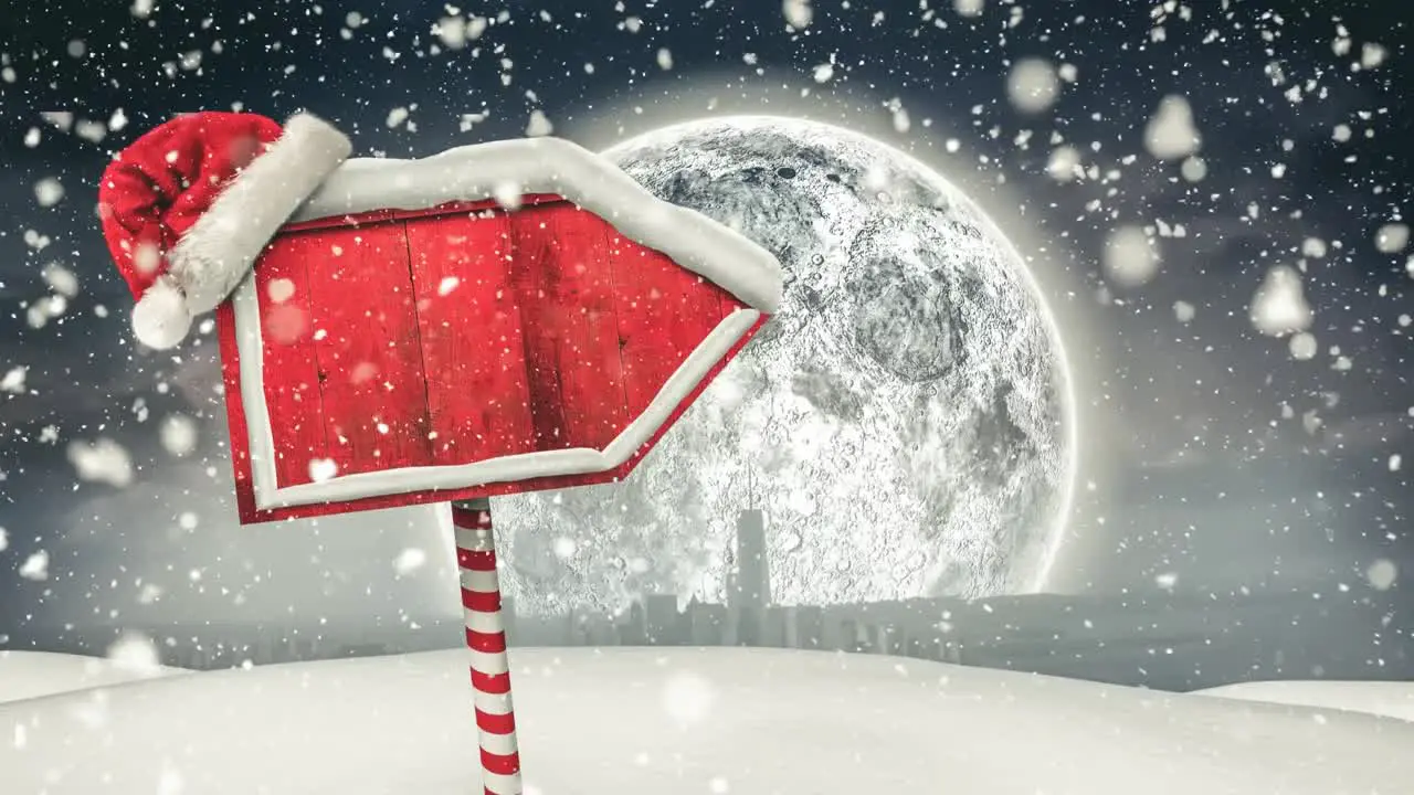 Snow falling and santa hat over red wooden sign post on winter landscape against moon in night sky