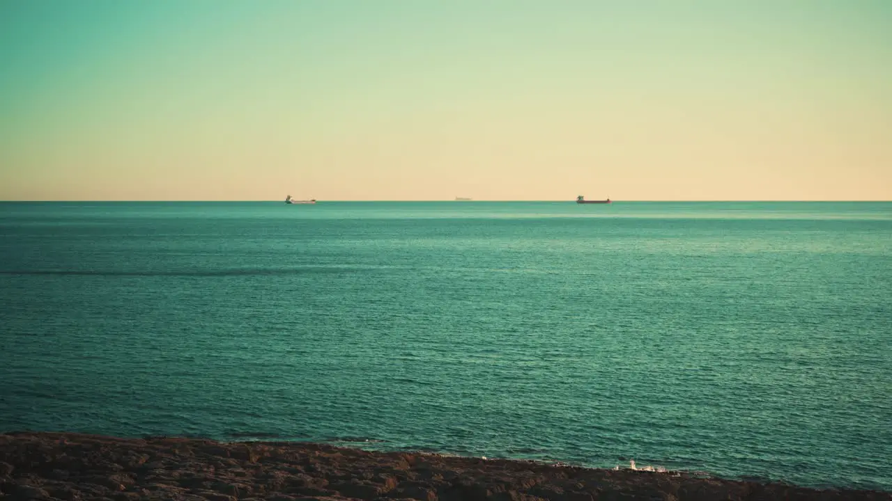 Ocean shoreline cliff rocks with cargo boats away at sunset with blue sky 4K