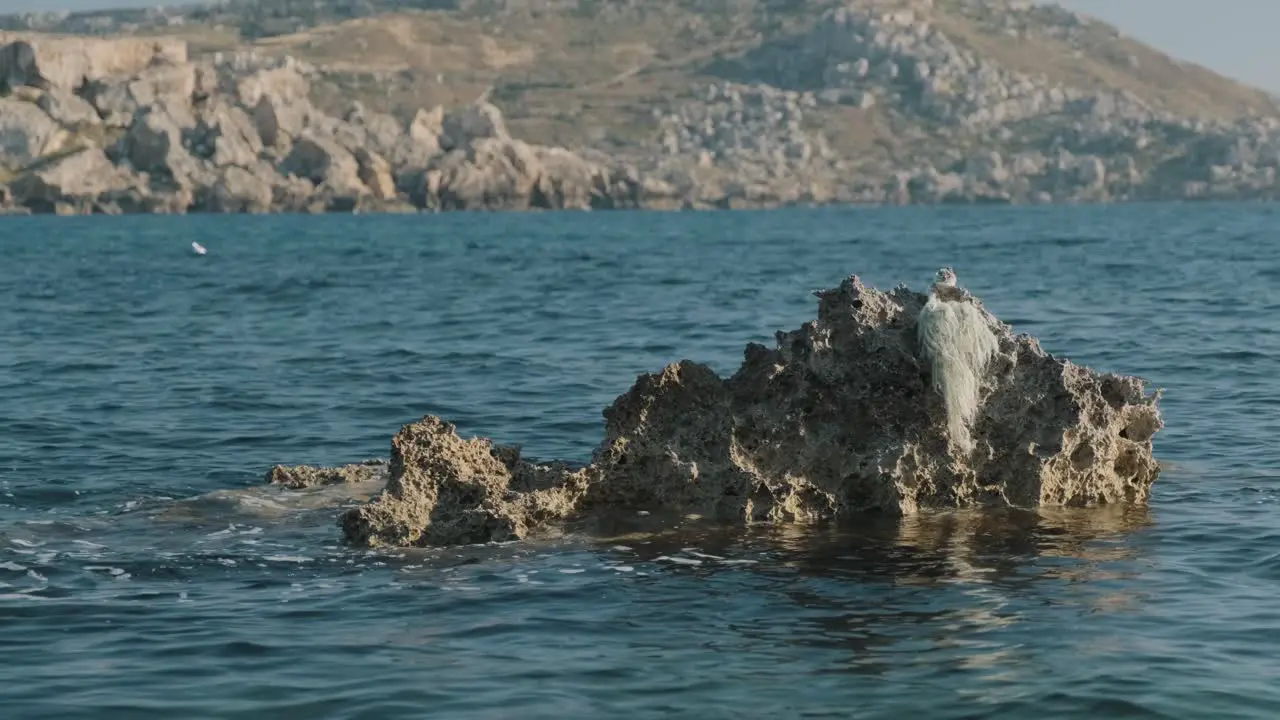 Water gently splashes on a rock formation in the middle of the sea with an arid stony mountain landscape in the background