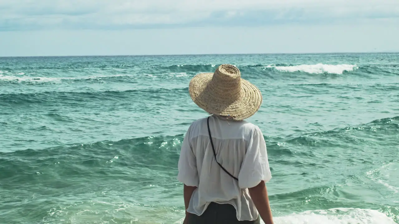 A woman with a big straw hat enjoys a walk by the sea