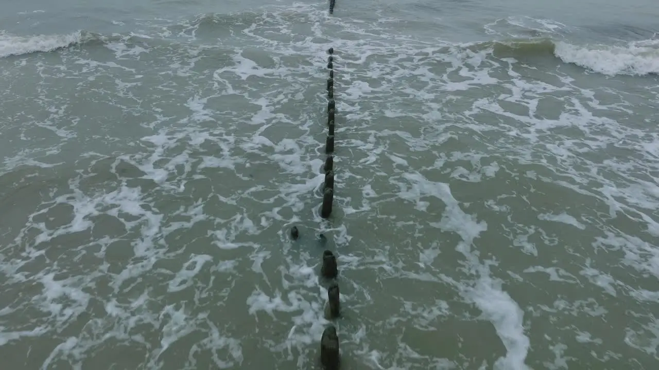 Aerial birdseye view of an old wooden pier at the Baltic sea coastline overcast winter day white sand beach wooden poles waves hitting shore wide drone shot moving forward low