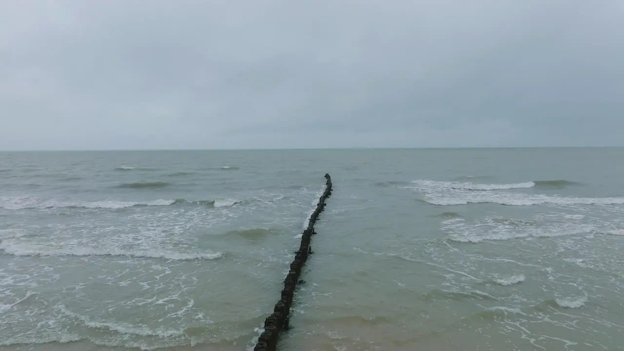 Aerial establishing view of an old wooden pier at the Baltic sea coastline overcast winter day white sand beach wooden poles waves hitting shore wide drone shot moving forward low