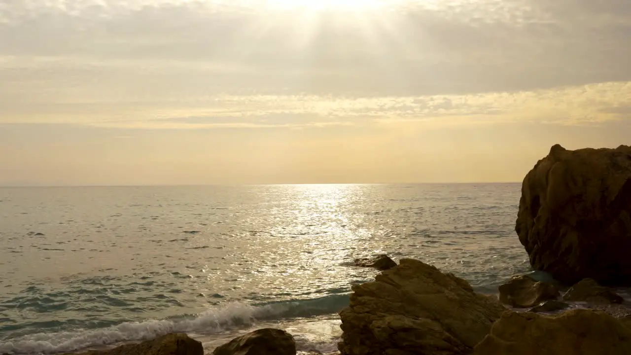 Sun rays flares through clouds on sky over sea water splashing rocky beach at twilight