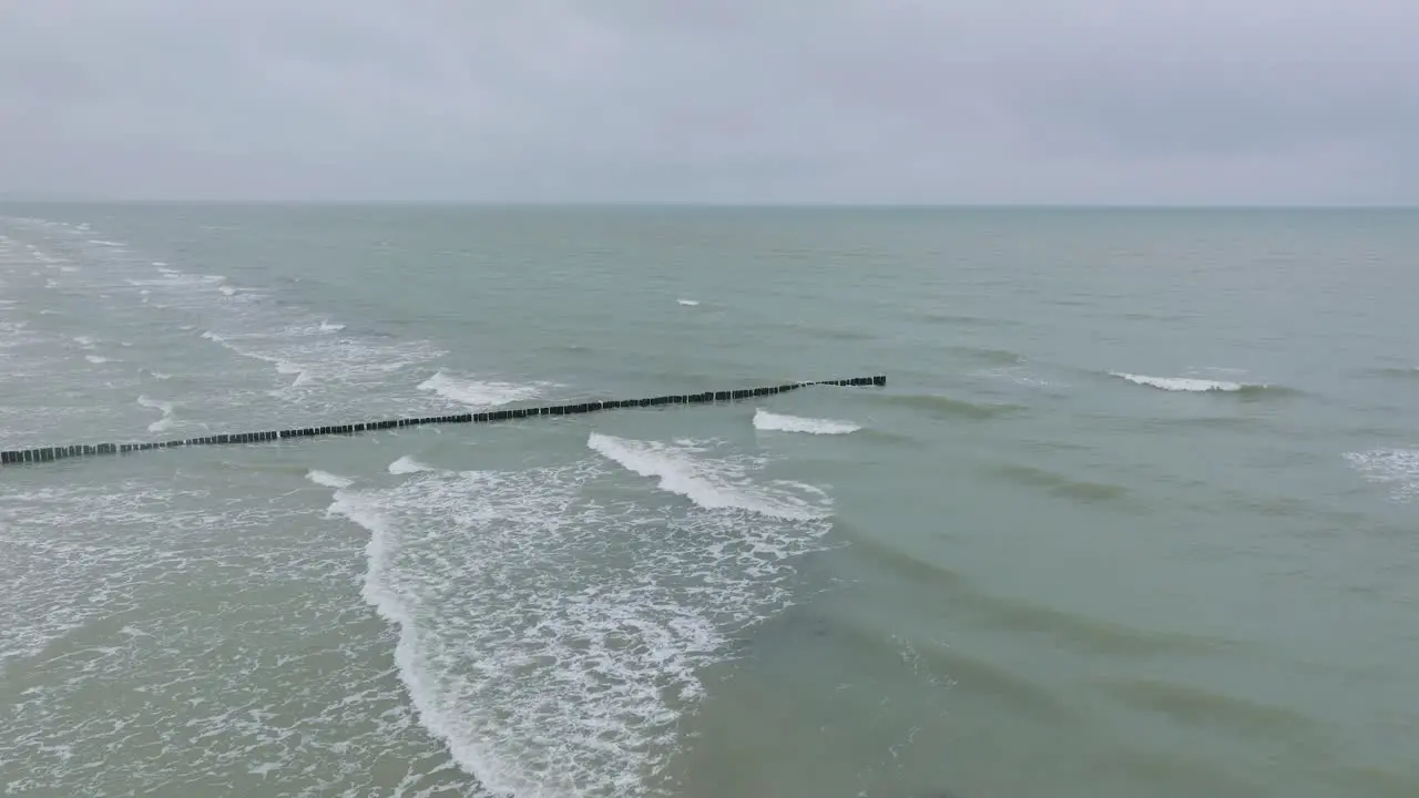 Aerial establishing view of an old wooden pier at the Baltic sea coastline overcast winter day white sand beach wooden poles waves hitting shore wide drone shot moving forward