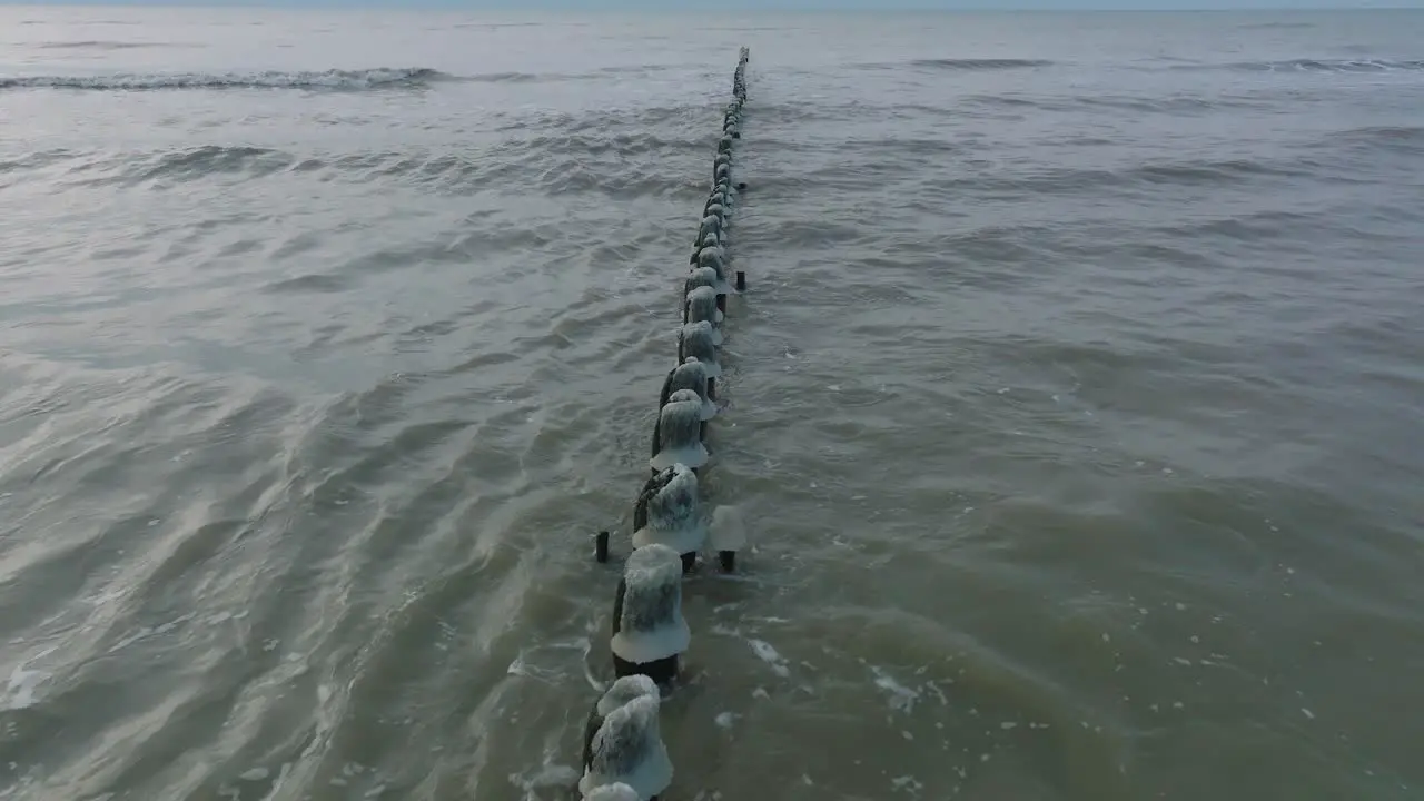 Aerial birdseye view of an old wooden pier at the Baltic sea coastline overcast winter day white sand beach covered in snow ice on wood poles calm seashore wide drone shot moving back low