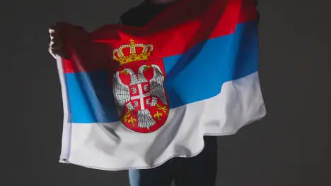 Studio Shot Of Anonymous Person Or Sports Fan Waving Flag Of Serbia Against Black Background