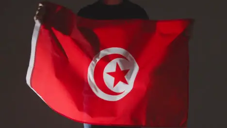 Studio Shot Of Anonymous Person Or Sports Fan Waving Flag Of Tunisia Against Black Background