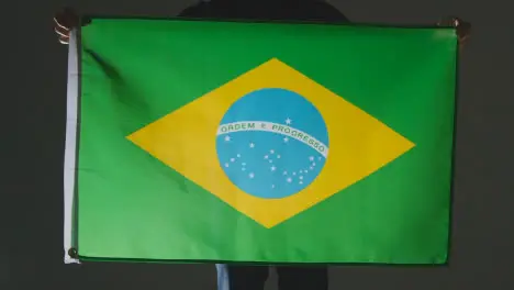 Studio Shot Of Anonymous Person Or Sports Fan Holding Flag Of Brazil Against Black Background