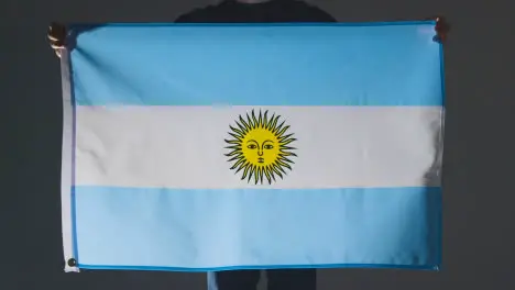 Studio Shot Of Anonymous Person Or Sports Fan Holding Flag Of Argentina Against Black Background