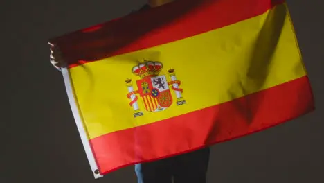 Studio Shot Of Anonymous Person Or Sports Fan Waving Flag Of Spain Against Black Background