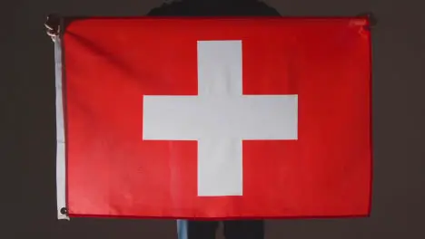 Studio Shot Of Anonymous Person Or Sports Fan Holding Flag Of Switzerland Against Black Background