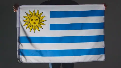 Studio Shot Of Anonymous Person Or Sports Fan Holding Flag Of Uruguay Against Black Background