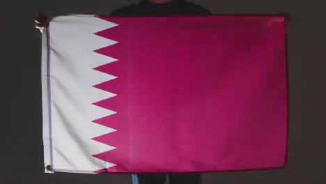 Studio Shot Of Anonymous Person Or Sports Fan Holding Flag Of Qatar Against Black Background