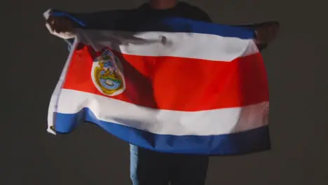 Studio Shot Of Anonymous Person Or Sports Fan Waving Flag Of Costa Rica Against Black Background