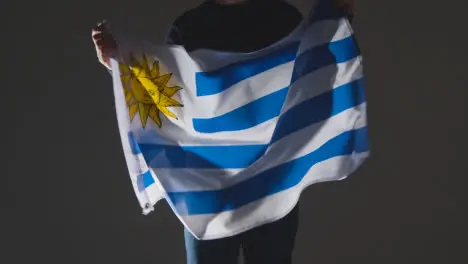 Studio Shot Of Anonymous Person Or Sports Fan Waving Flag Of Uruguay Against Black Background