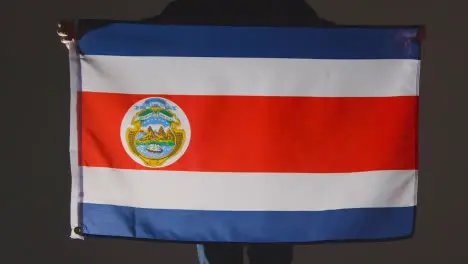 Studio Shot Of Anonymous Person Or Sports Fan Holding Flag Of Costa Rica Against Black Background