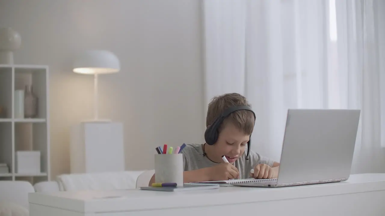 schooler boy is viewing learning video by notebook and listening teacher by headphones drawing in copybook