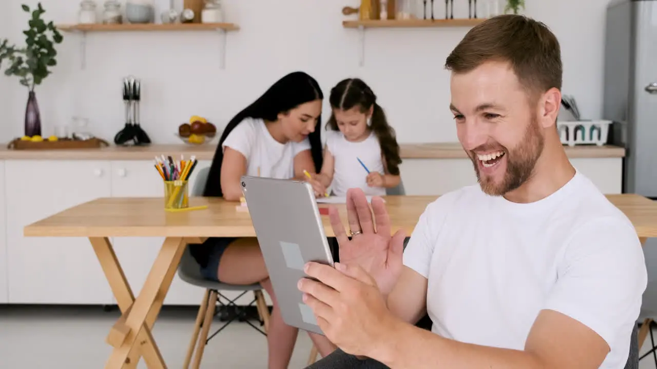 Happy Handsome Man Having A Video Call Using A Digital Tablet At Home While On Background His Wife And Little Daughter Sitting At Table And Drawing Together