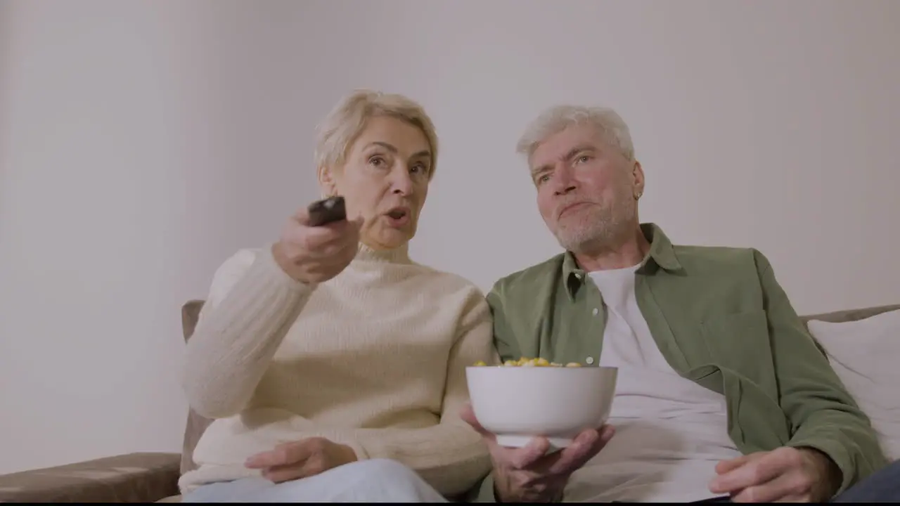 Happy Senior Couple Eating Caramel Popcorn And Watching Tv While Sitting On Sofa At Home