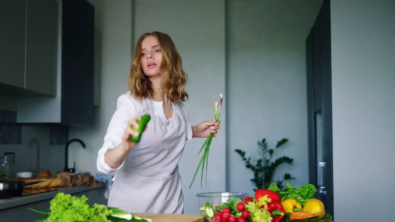 Happy girl dancing and singing in kitchen using cucumber as microphone