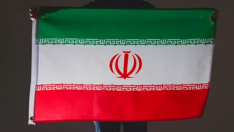 Studio Shot Of Anonymous Person Or Sports Fan Holding Flag Of Iran Against Black Background