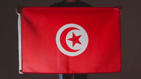 Studio Shot Of Anonymous Person Or Sports Fan Holding Flag Of Tunisia Against Black Background