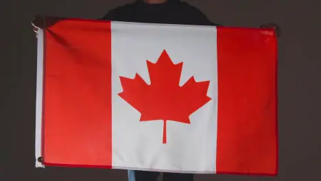 Studio Shot Of Anonymous Person Or Sports Fan Holding Flag Of Canada Against Black Background