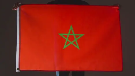 Studio Shot Of Anonymous Person Or Sports Fan Holding Flag Of Morocco Against Black Background