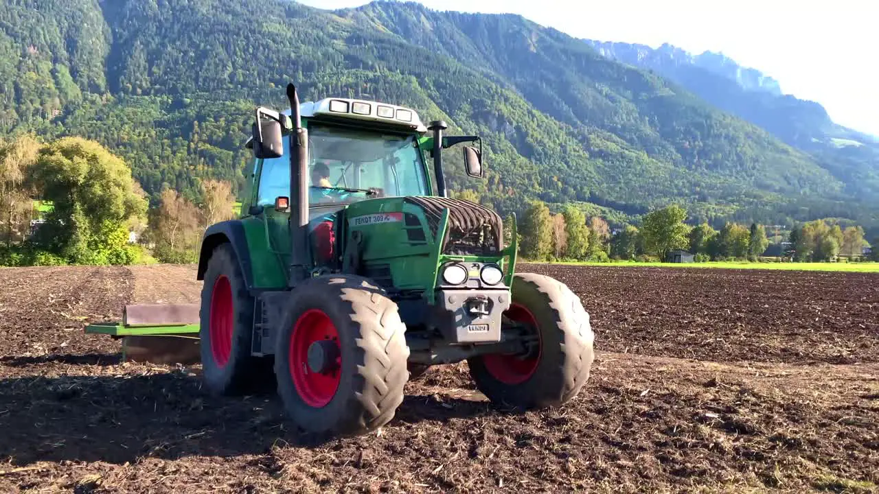 A four wheeler tractor with the driver flattening farm lands with mountain and green trees in the background