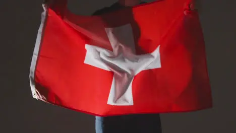 Studio Shot Of Anonymous Person Or Sports Fan Waving Flag Of Switzerland Against Black Background