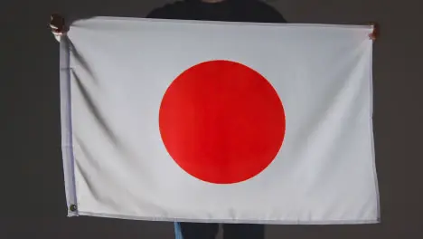 Studio Shot Of Anonymous Person Or Sports Fan Holding Flag Of Japan Against Black Background