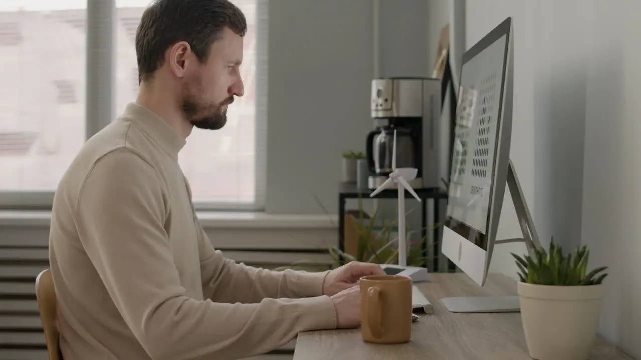 Side View Of Man Working With Pc And Drinking Coffee Sitting At Desk In The Office