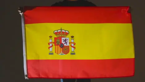 Studio Shot Of Anonymous Person Or Sports Fan Holding Flag Of Spain Against Black Background