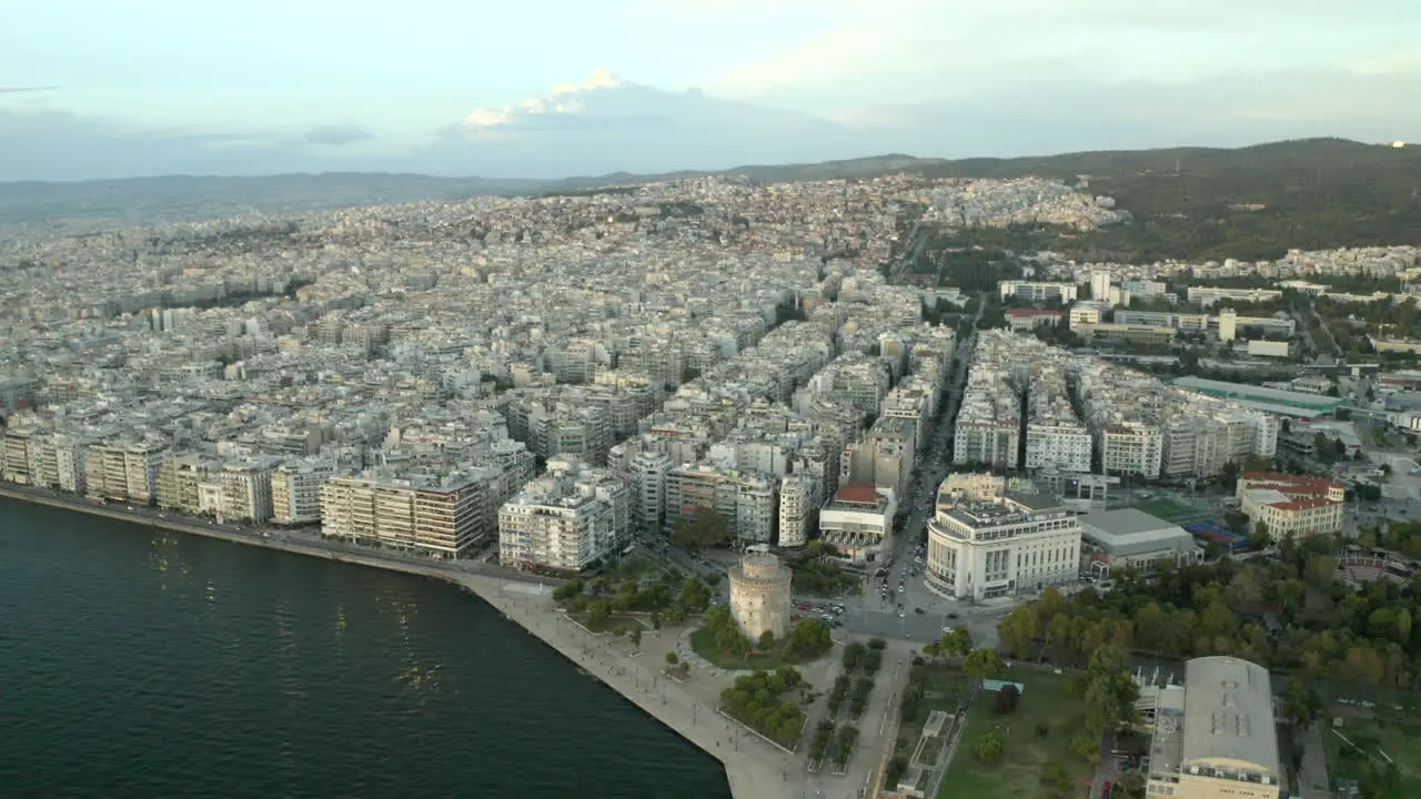 cinematic wide drone aerial shot of northern Greece city of Thessaloniki with the sea and city and white tower in view at sunset moving backwards in 4k