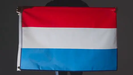 Studio Shot Of Anonymous Person Or Sports Fan Holding Flag Of Netherlands Against Black Background