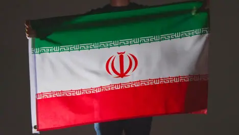 Studio Shot Of Anonymous Person Or Sports Fan Waving Flag Of Iran Against Black Background