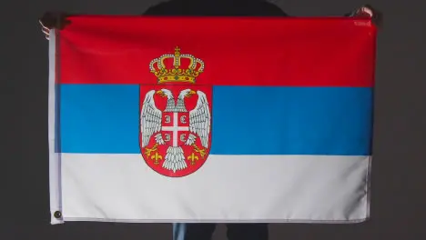 Studio Shot Of Anonymous Person Or Sports Fan Holding Flag Of Serbia Against Black Background