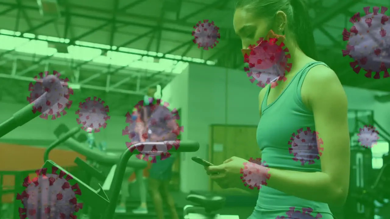 Covid 19 cells floating against woman wearing face mask using smartphone at the gym