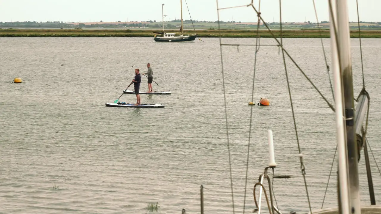 Footage showing two men standup paddle boarding across the frame this is filmed at Heybridge basin in Essex