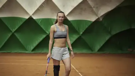 Young long haired woman in sportswear is walking on the indoor tennis court with racket in her hand Green tent behind Slow motion