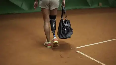 Tracking footage of a invalid athlete walking on the tennis court with the racket in her bag Back view Slow motion