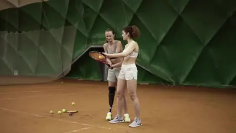 A beautiful female coach with a prosthesis on her leg shows her ward the nuances of the ball hitting in tennis standing behind her Sports girls