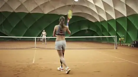 Backside footage of two athletic girls playing tennis in a covered tennis court Woman in the frame with a leg prosthesis