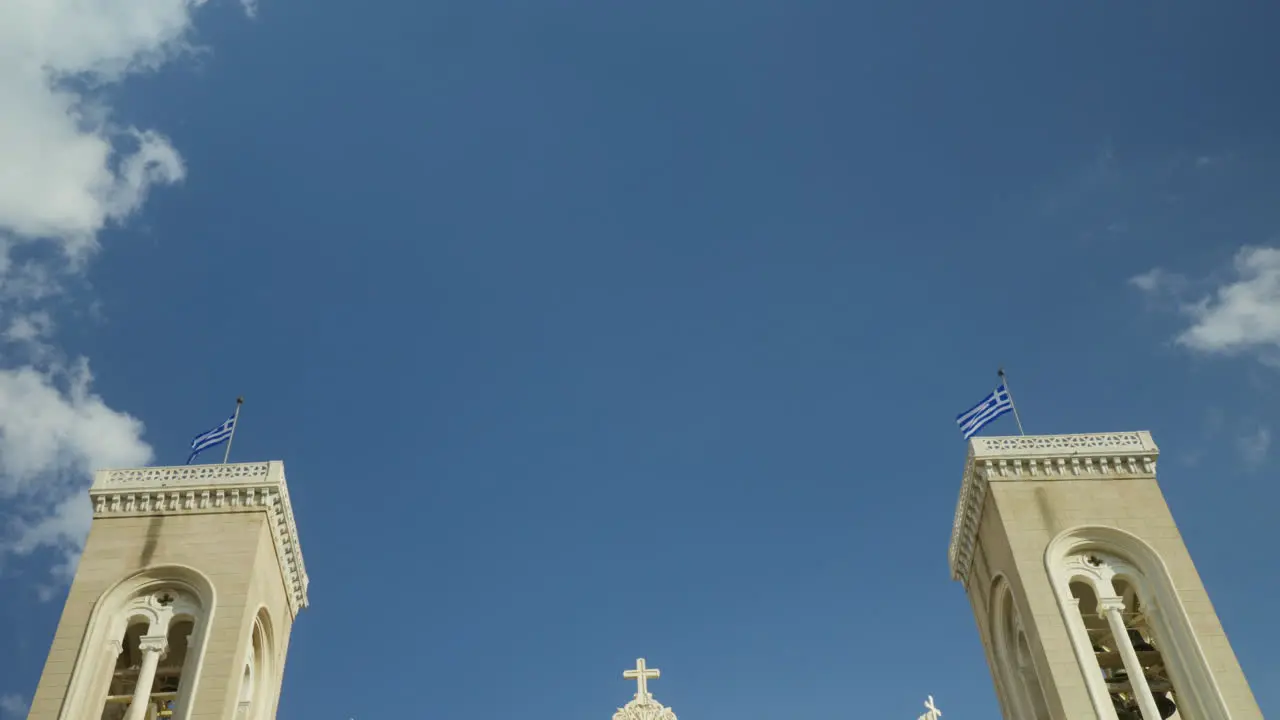 Twin bell towers against a vibrant blue sky with Greek flags