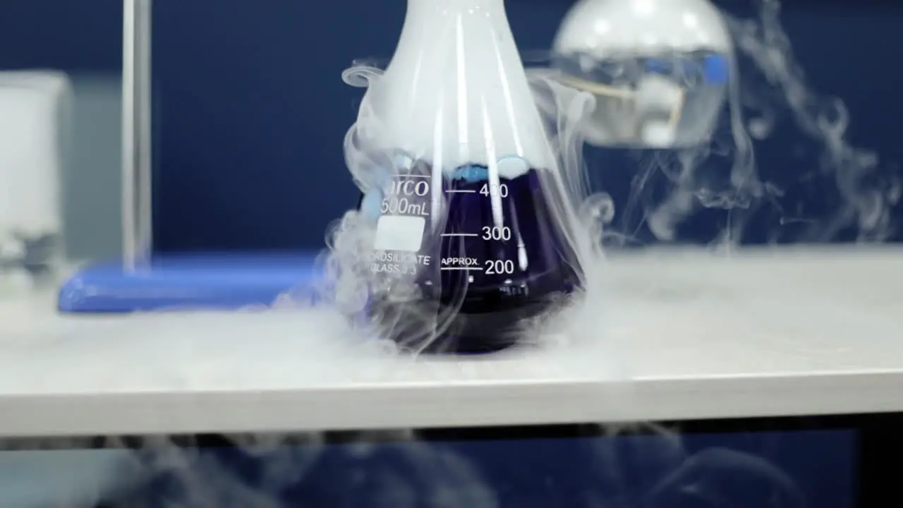 In detailed slow-motion a glass beaker contains a vibrant blue liquid undergoing a chemical reaction as white smoke gracefully emanates illustrating the dynamics of scientific exploration